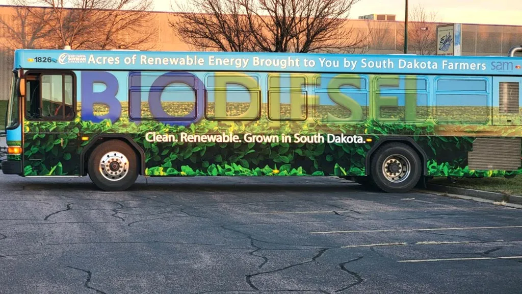 A bus with an advertisement for biodiesel featuring large colorful text reading 'BIODIESEL' over a background of green plants, with text above saying 'Acres of Renewable Energy Brought to You by South Dakota Farmers' and below stating 'Clean. Renewable. Grown in South Dakota.'