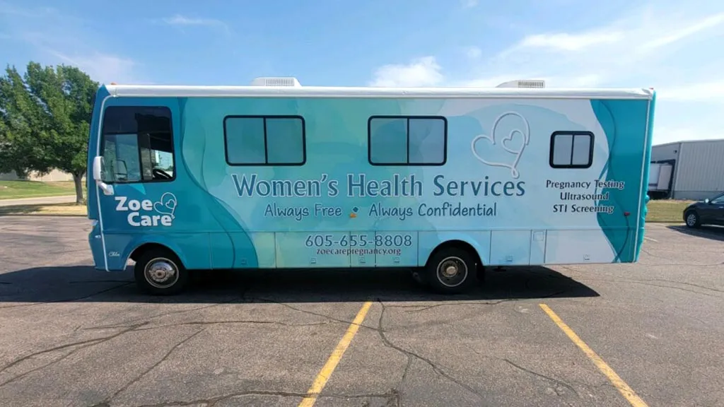 Side view of a teal and white mobile clinic bus for Zoe Care offering Women's Health Services with text stating Always Free, Always Confidential, and phone number 605-655-8808, listing services including Pregnancy Testing, Ultrasound, and STI Screening.