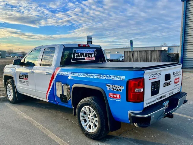 White and blue pickup truck parked near industrial buildings with company branding for Lambert Heating, advertising air conditioning and HVAC services, with contact phone number visible.
