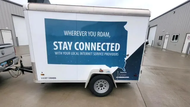White utility trailer with a blue outline of a state and the text: 'WHEREVER YOU ROAM, STAY CONNECTED WITH YOUR LOCAL INTERNET SERVICE PROVIDERS.' The trailer is parked on a wet concrete surface between two industrial buildings.