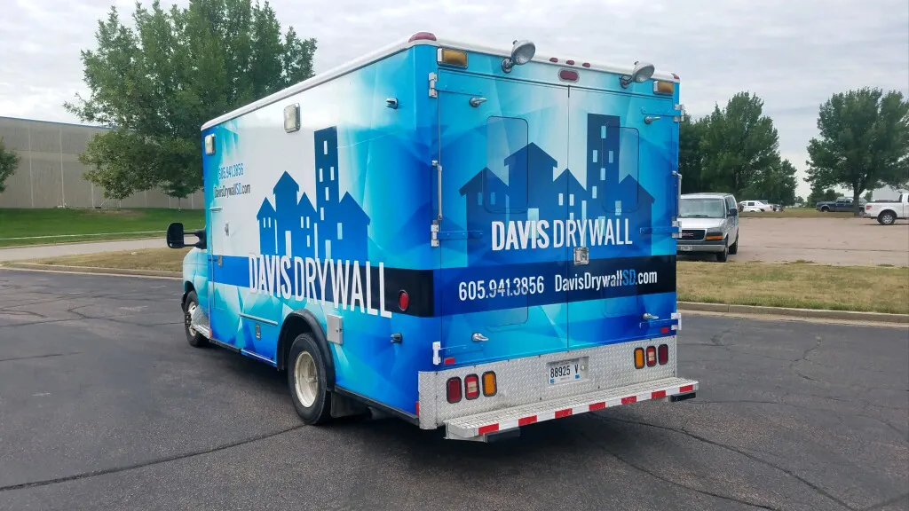 Blue delivery truck with 'Davis Drywall' and a city skyline graphic printed on its sides and back, parked on an asphalt surface with trees and other vehicles in the background.