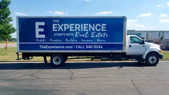 White box truck with a blue advertisement panel reading 'The Experience Starts With Real Estate' and listing services including rentals, furniture, builders, insurance, movers along with the website TheExperience.com and phone number 940-5544.