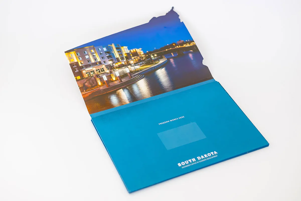 Open folder shaped like the state of South Dakota, featuring a night cityscape photo on the inner top flap and text 'Freedom Works Here' and 'South Dakota' on the bottom flap.