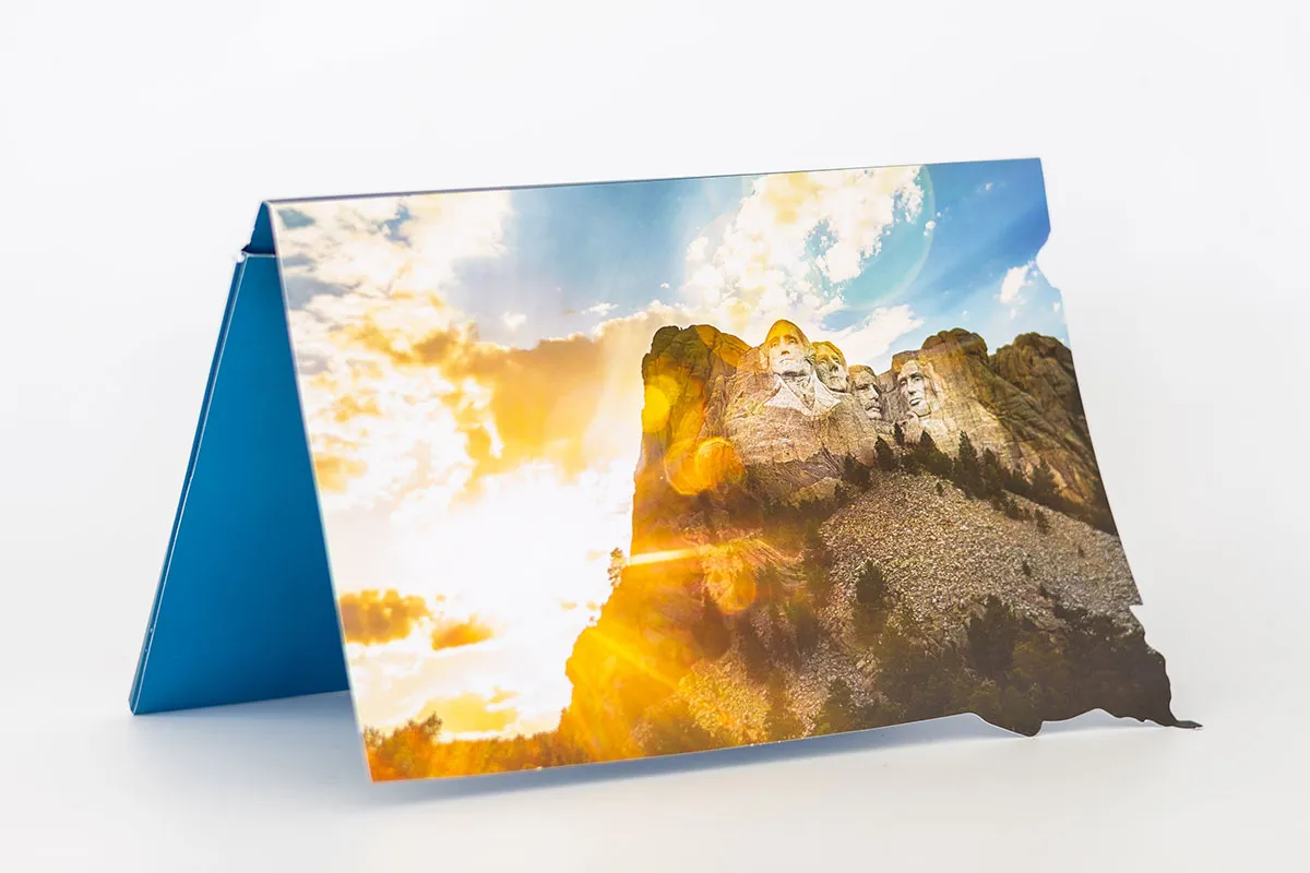 Folded card shaped like South Dakota displaying Mount Rushmore with sun rays and partly cloudy sky in the background.