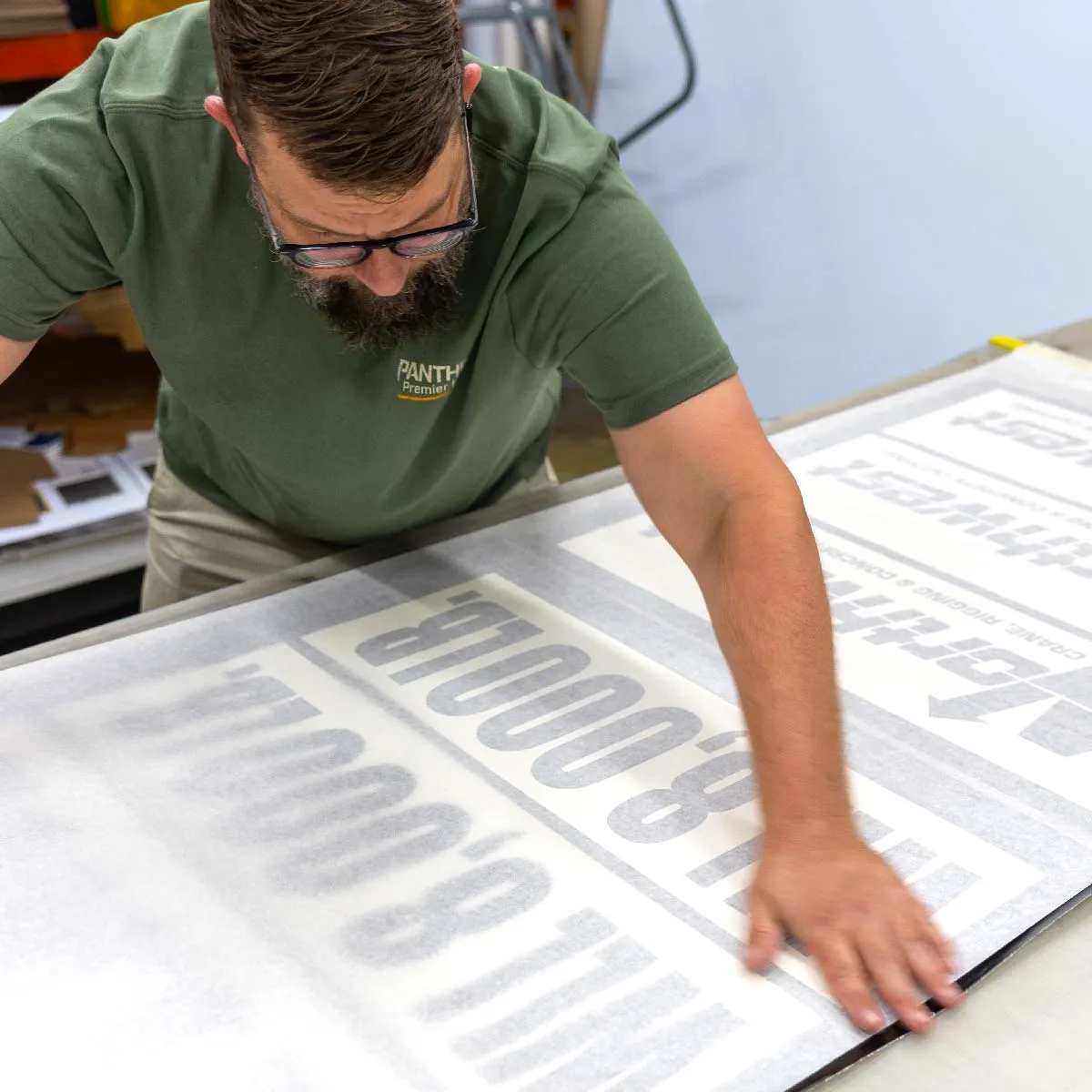 Man wearing glasses and a green shirt with text working on a large sheet that has printed words partially visible through a semi-transparent material.