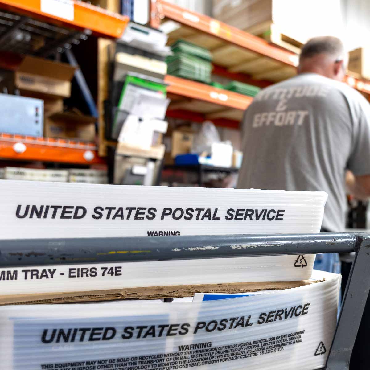 Close-up of two United States Postal Service plastic trays on a metal cart inside a warehouse with shelves and boxes in the background, and a man wearing a gray shirt with the words 'ATTITUDE & EFFORT' on the back, working in the background.