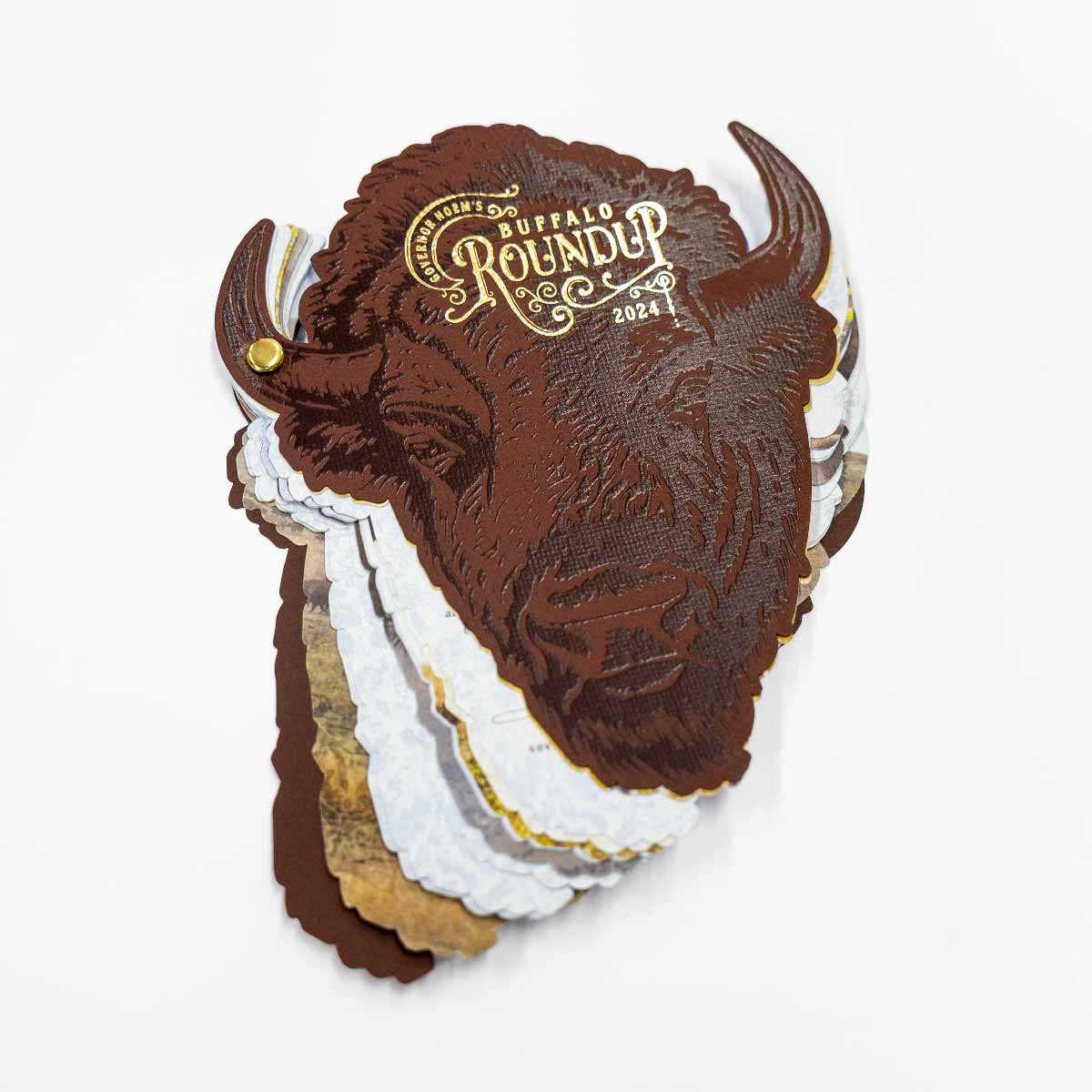 Stack of buff-colored cards shaped like a buffalo head with embossed details and gold text reading "Governor Noem's Buffalo Roundup 2024", held together by a brass fastener on a white background.