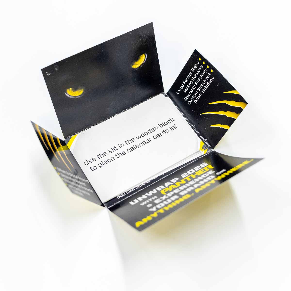 Open black printed cardboard packaging with yellow panther eyes on the inner flap and text instructions inside reading 'Use the slit in the wooden block to place the calendar cards in!' with additional printed information about large format signs and mailing services on the flaps.