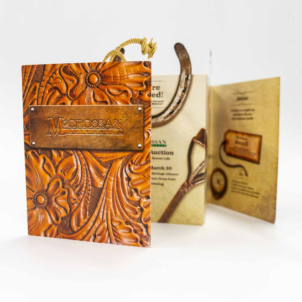 A folded brochure with a brown embossed floral leather design and a metal plate that reads 'McCrossan Boys Ranch'; the inside pages display vintage-style images of a horseshoe and a shovel with text about an auction event.