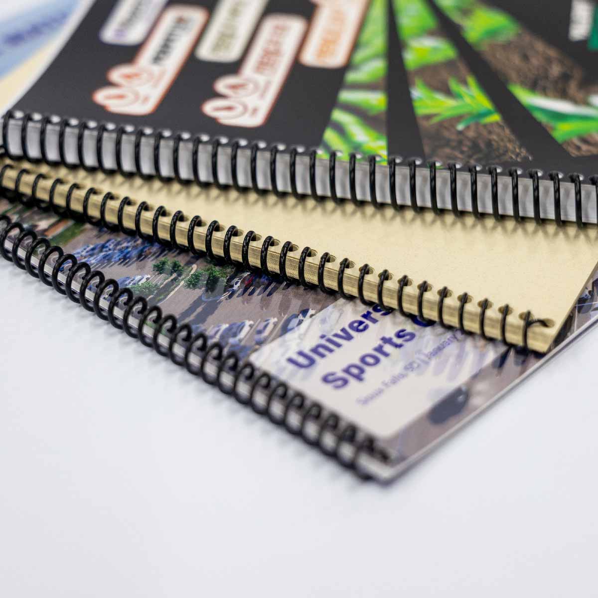 Close-up of three spiral-bound notebooks stacked on a white surface, featuring varied cover designs including a labeled sports-themed cover and a plain beige cover.