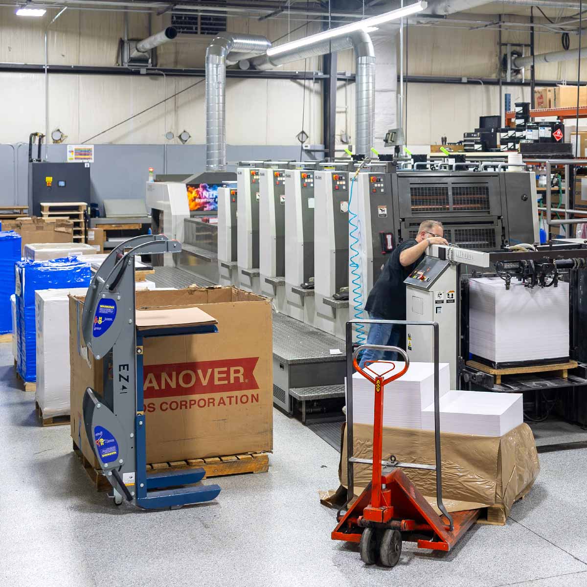 Worker operating a large commercial printing press in an industrial printing facility, with stacks of paper and pallets nearby, along with a red pallet jack and printing supplies.
