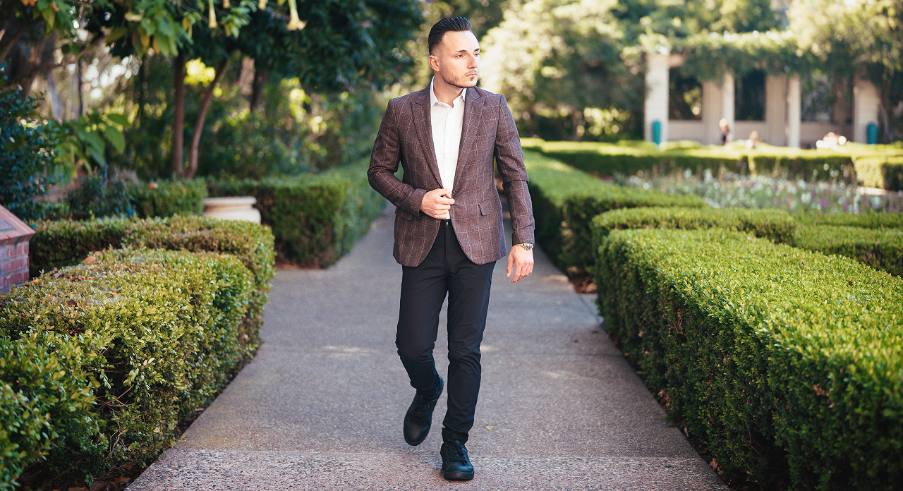 Man in a brown jacket and black pants walking on a garden path bordered by trimmed hedges.