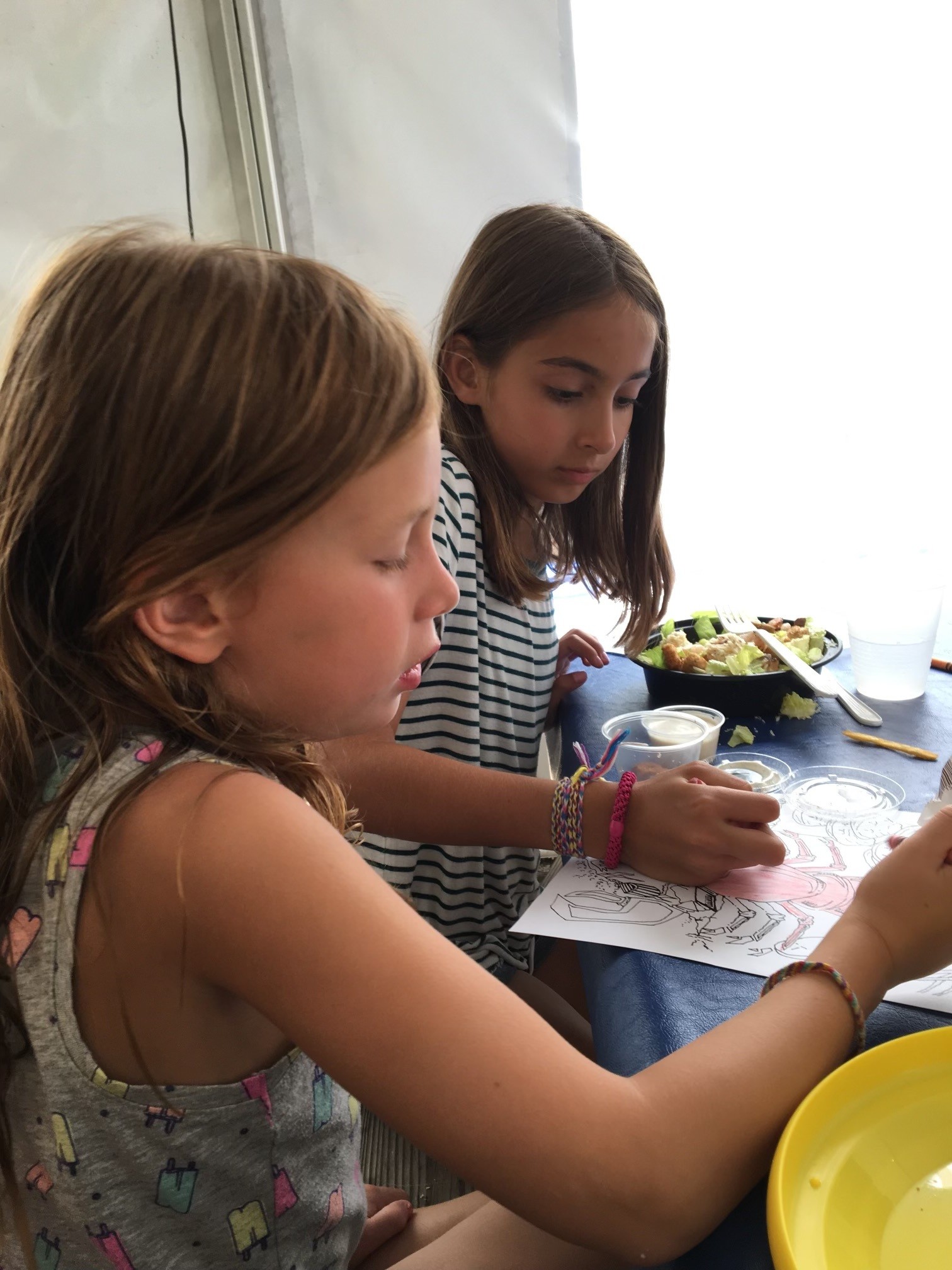 Two young girls sitting at a table, one coloring on a paper with a pink crayon while the other watches, with a bowl of salad and a glass of water nearby.