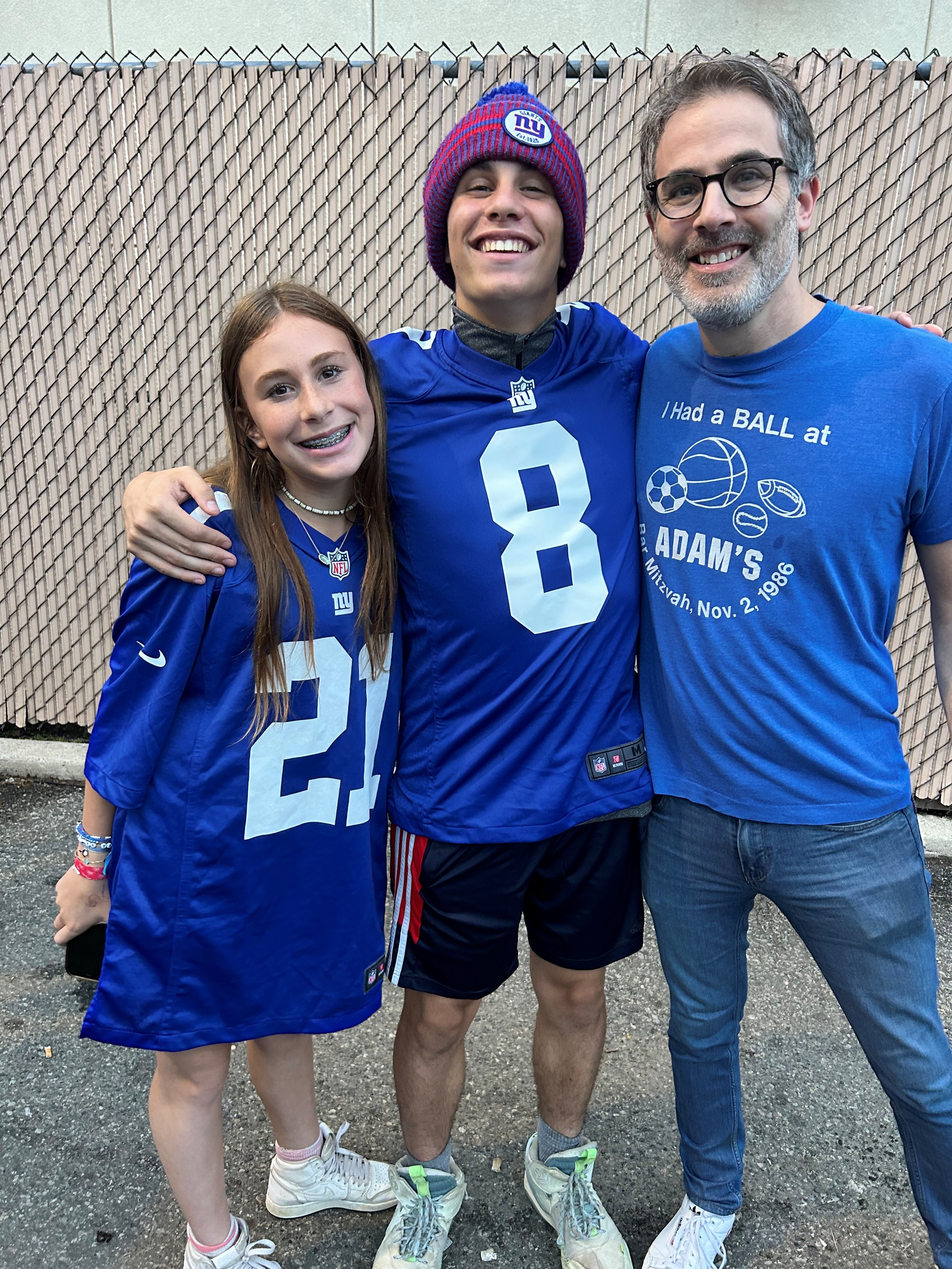 Three smiling people standing closely in front of a chain-link fence: a girl wearing a blue jersey with number 21 and white sneakers, a boy in a blue jersey with number 8, red and blue knit hat, and white sneakers, and a man with glasses, a beard, and a blue t-shirt with sports balls and text, wearing jeans and white shoes.