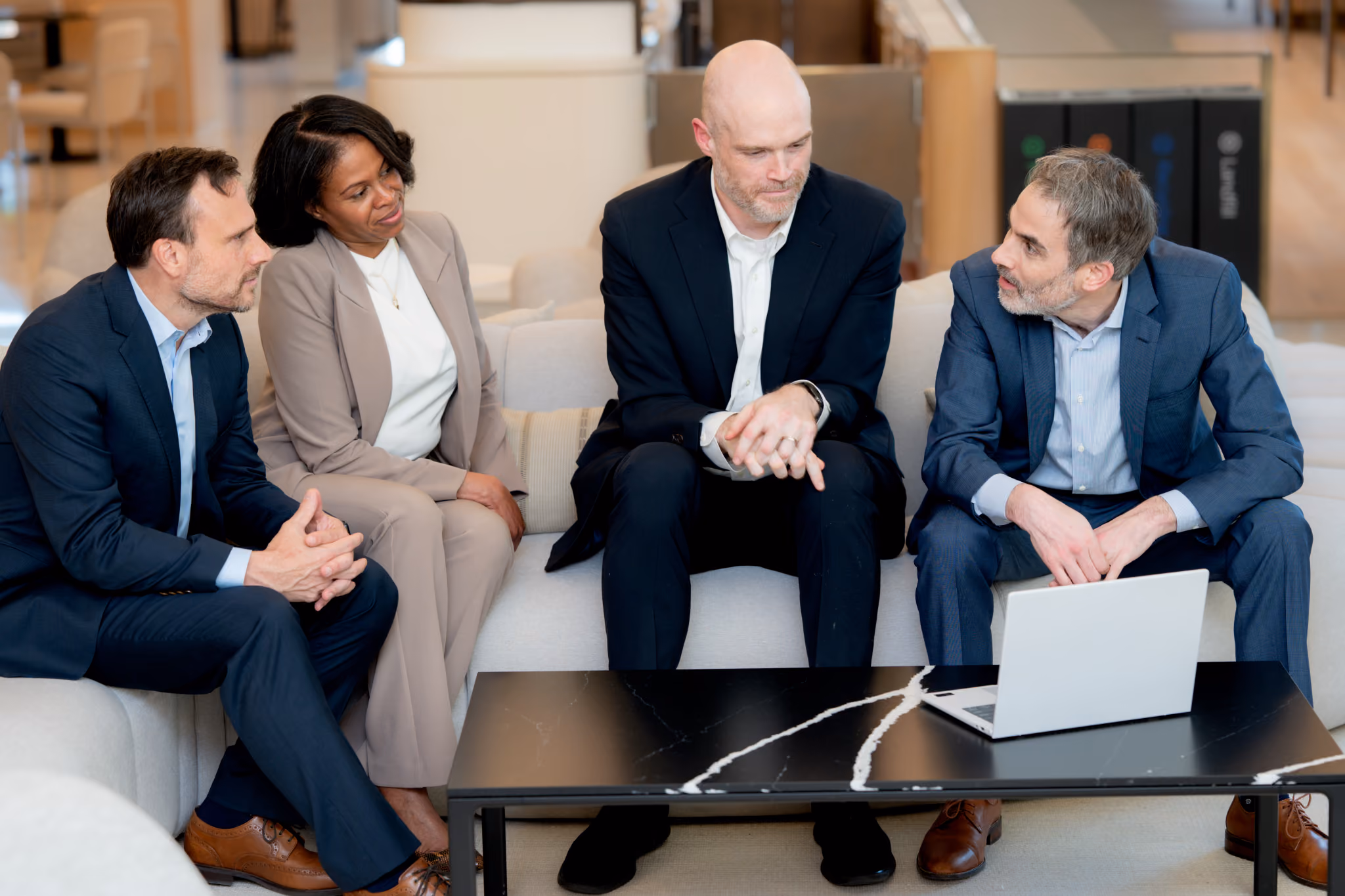Four Kohn Dumain colleagues sitting on a light gray sofa in a modern office discussing a laptop on a black marble coffee table.