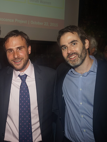 Two men in business attire smiling indoors, one wearing a suit with a purple tie and the other in a blue shirt without a tie, with a partially visible projection screen in the background.