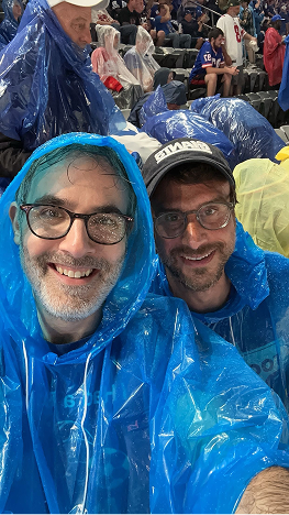Two men wearing blue rain ponchos and glasses smiling at the camera in an outdoor stadium with other people in rain ponchos in the background.