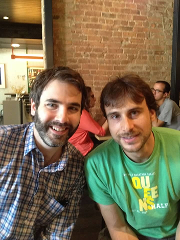Two men smiling at the camera sitting indoors against a brick wall; one wears a checkered shirt and the other a green t-shirt with yellow and white text.