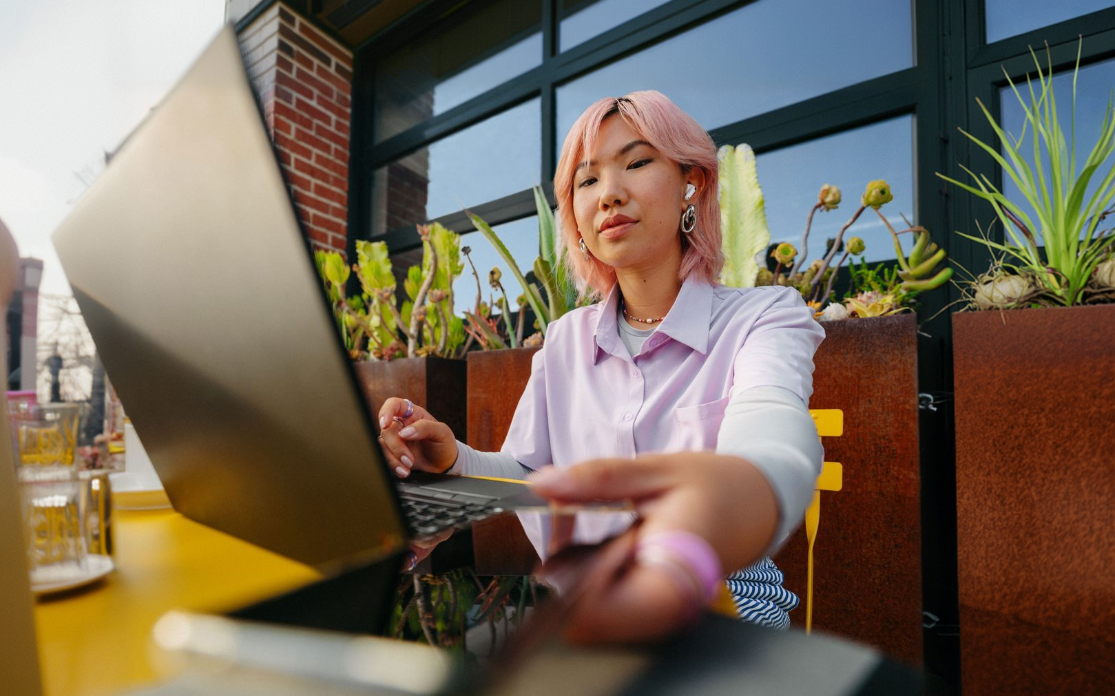 Person with pink hair handing a credit card toward a laptop at an outdoor café table.