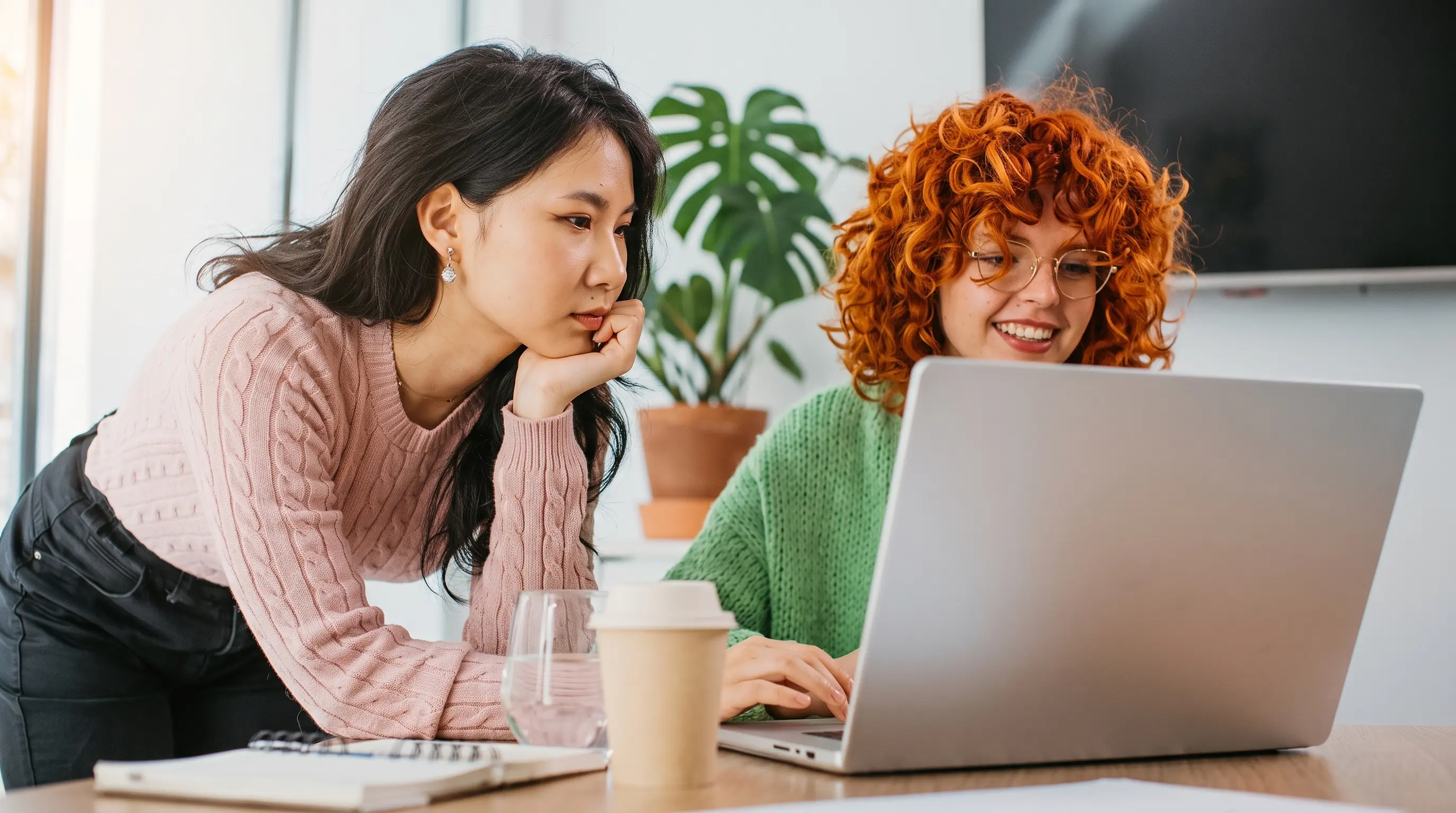 Two young women working together at a laptop on a table with a notebook, cup, and glass.