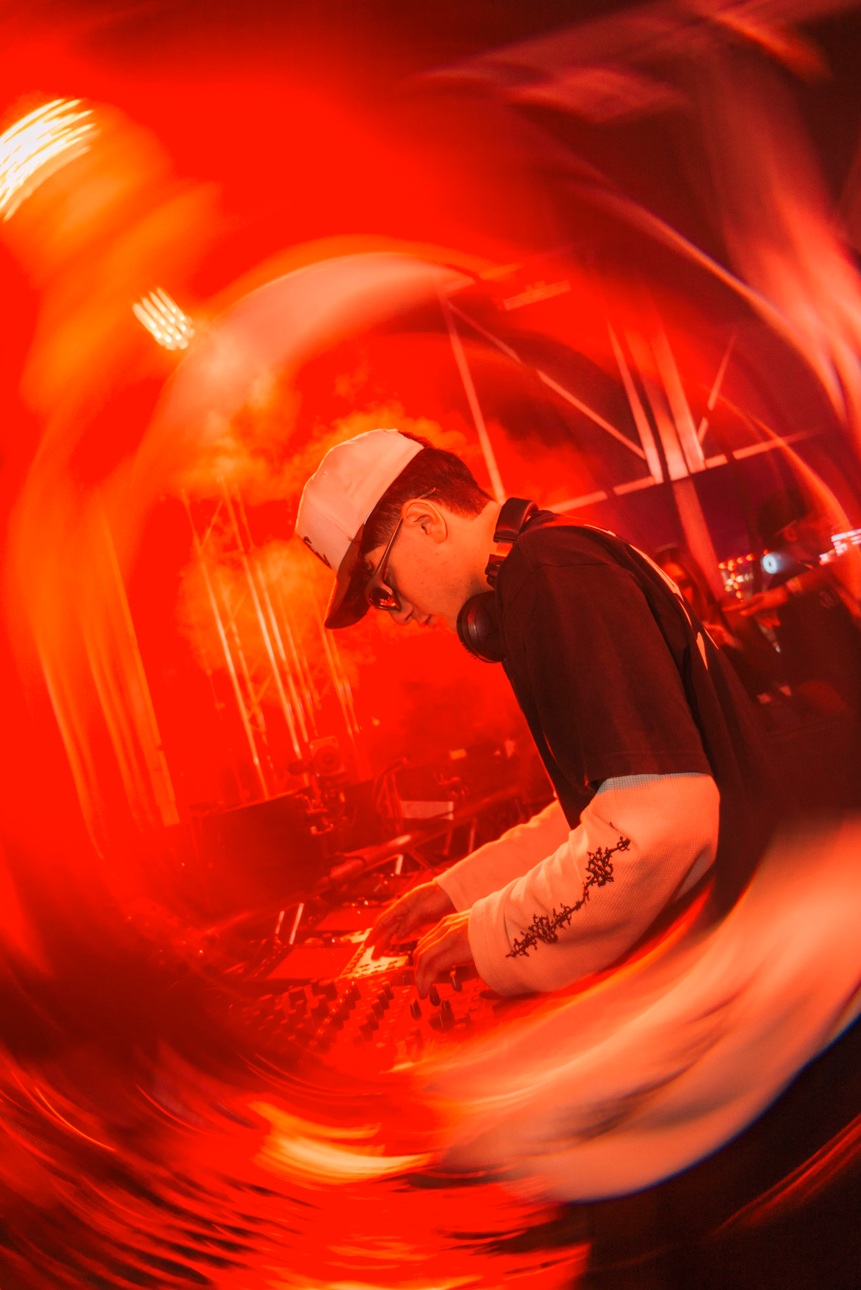 DJ wearing a cap and headphones mixing music on a console in red concert lighting.