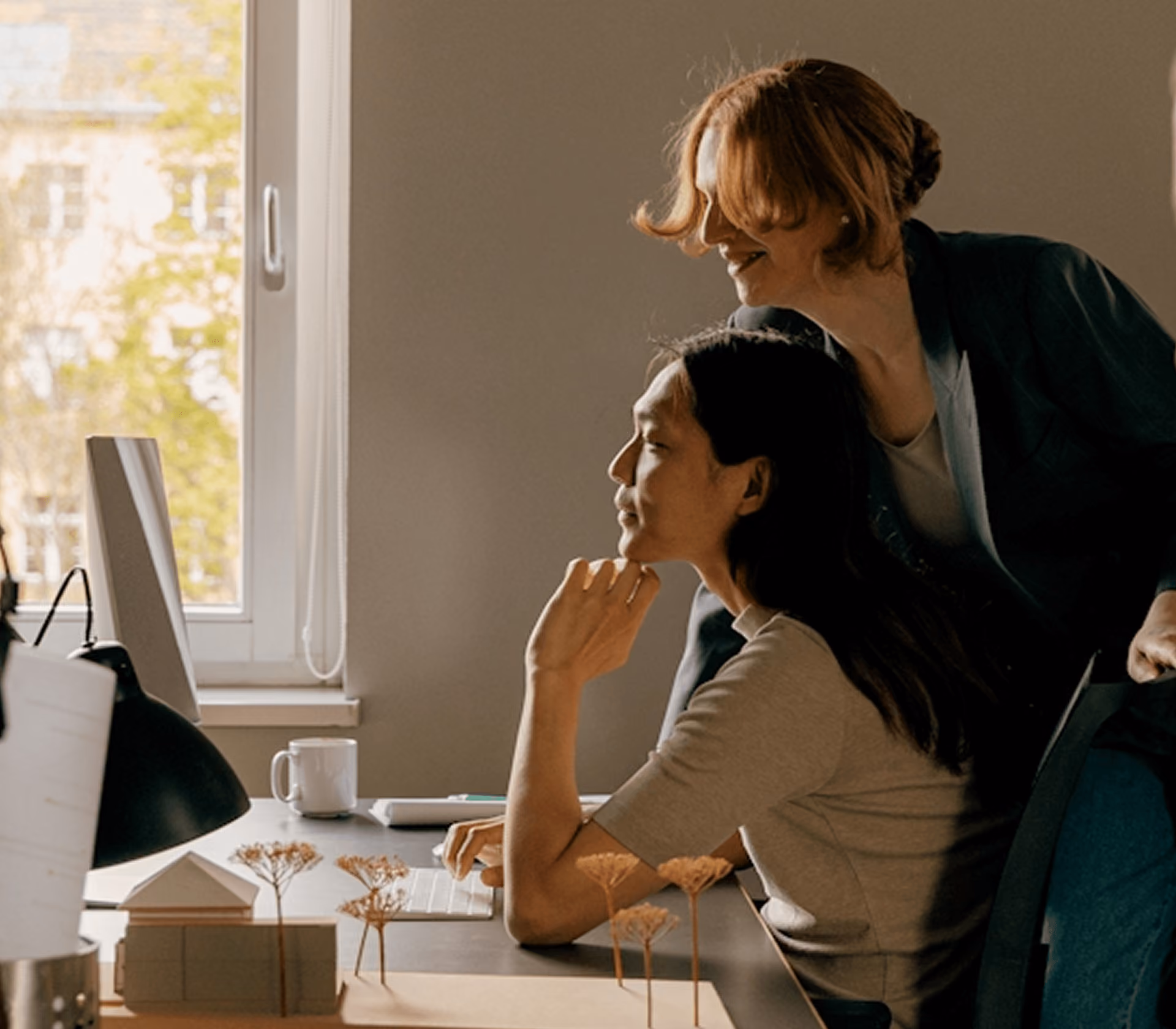 Two women working together at a computer in a warmly lit office with a window view.