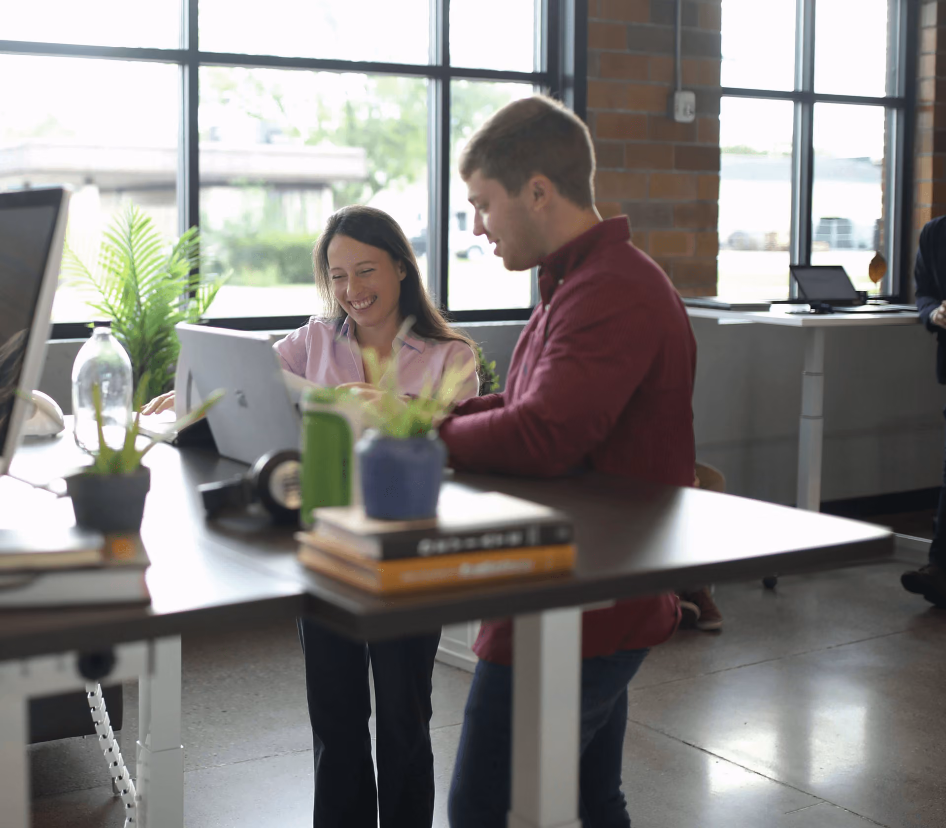 Two coworkers smiling and discussing while using a tablet at a shared office desk by large windows.