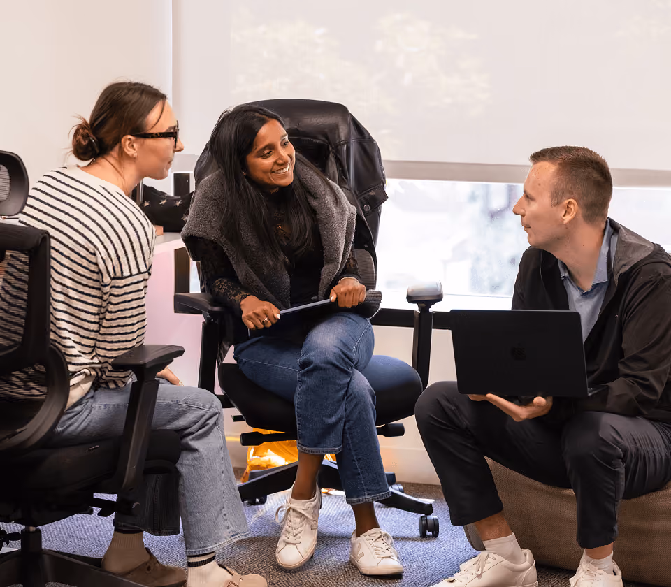 Three people seated indoors in a casual meeting; one holds a laptop, another a tablet.
