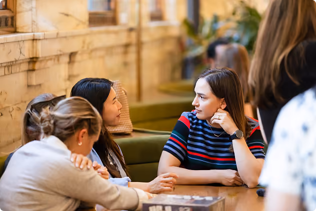 Two women engaged in deep conversation at a table in a cozy indoor setting with others nearby.