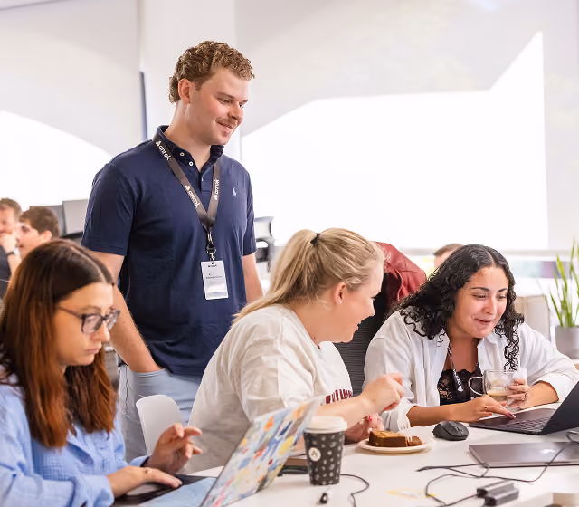 Four coworkers collaborate in a bright office, one standing and three seated, using laptops and sharing food.