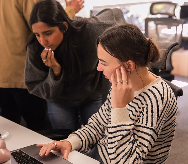 Two women working together at a desk, focused on a laptop screen in an office setting.