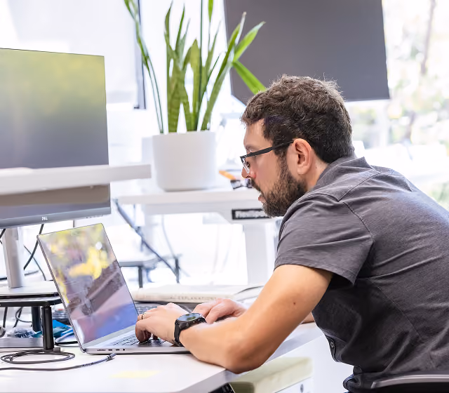 Man wearing glasses working on a laptop at a desk with monitors and a potted plant nearby.