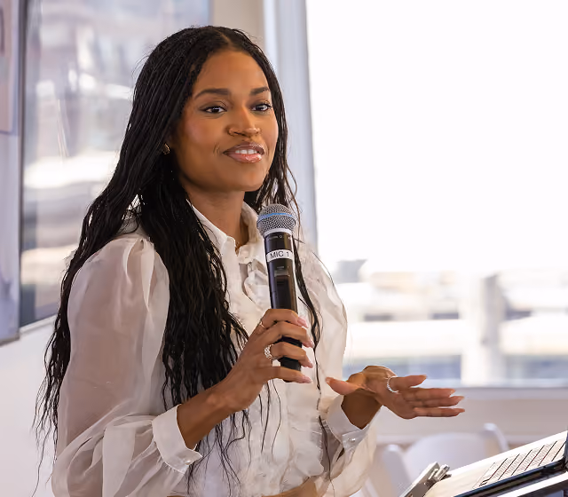Woman with long hair speaking into a microphone during a presentation.
