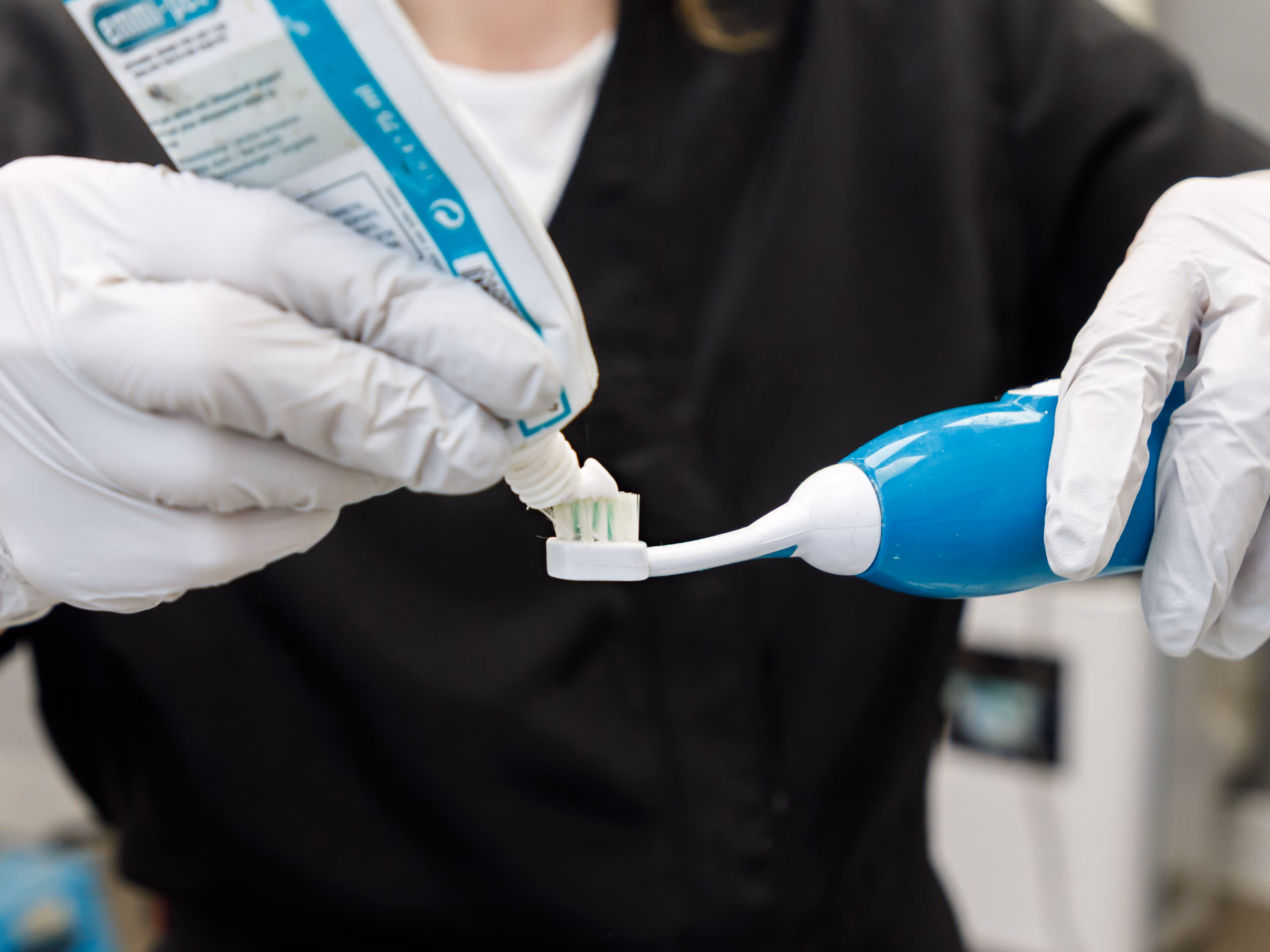 Person wearing gloves squeezing toothpaste onto a blue electric toothbrush head.