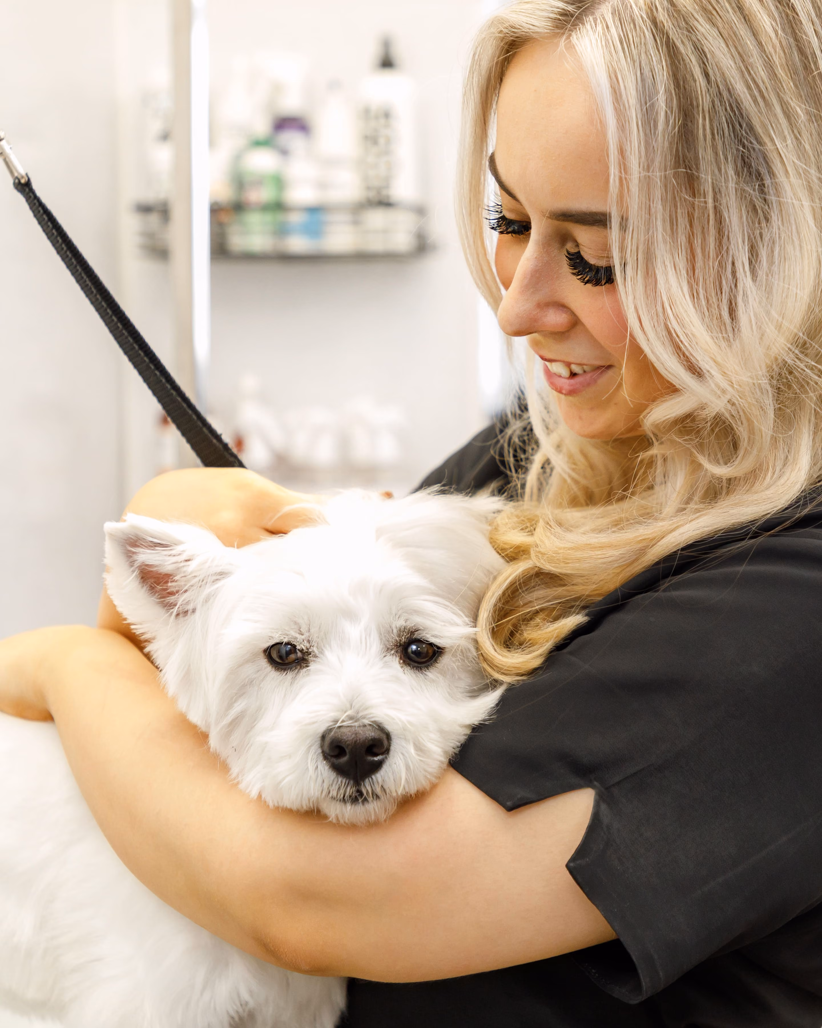 Smiling groomer with long blonde hair hugging a white dog during grooming session.