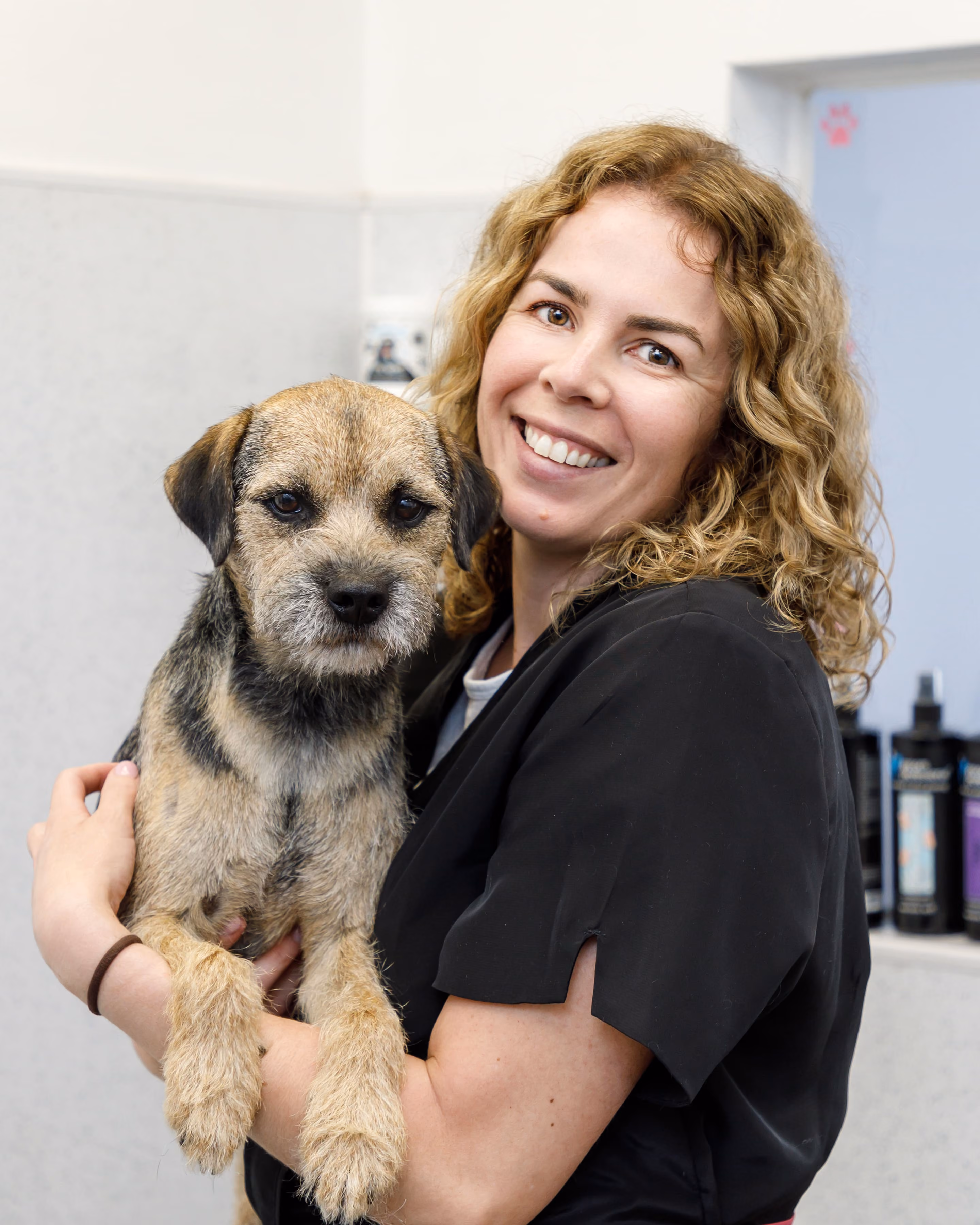 Woman with curly hair holding a small brown and black dog in a grooming setting.