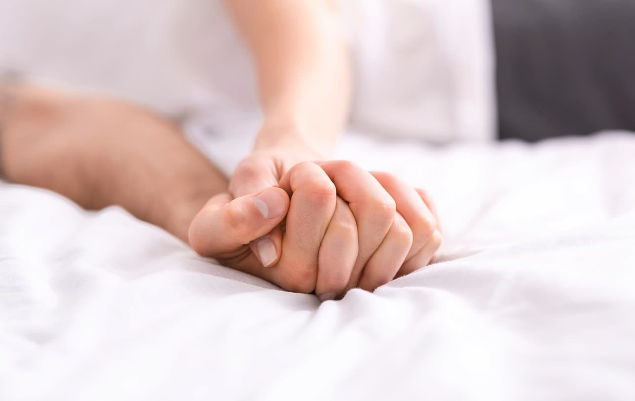 A man and woman sit on a bed holding hands, conveying intimacy, connection, and affection.