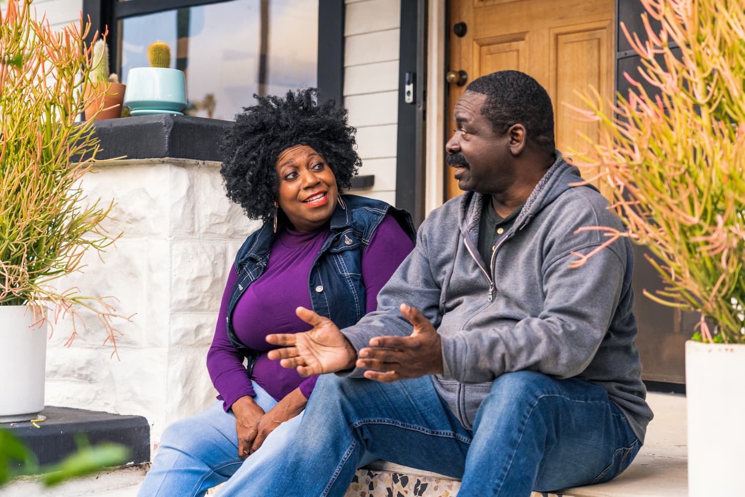 A man and woman sit on house steps, sharing a relaxed moment of conversation together.