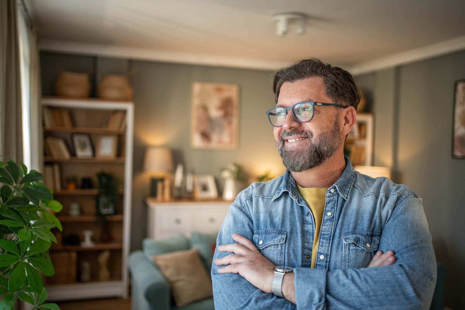 A man with glasses and a beard stands in a cozy living room, surrounded by furniture and decor.