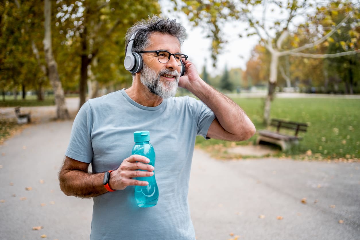 Bearded older man wearing headphones talks on his phone while walking through a green park.