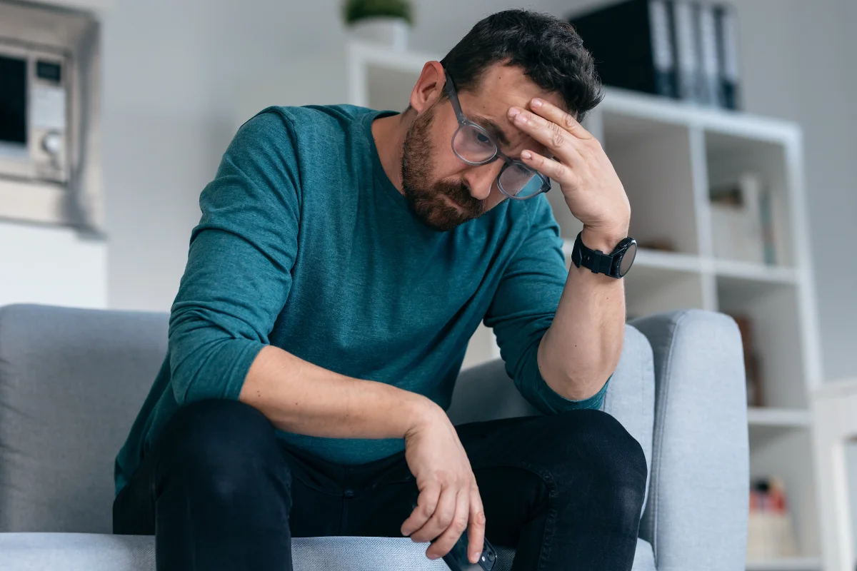 Man sitting on couch holding head showing stress and erectile dysfunction concerns.