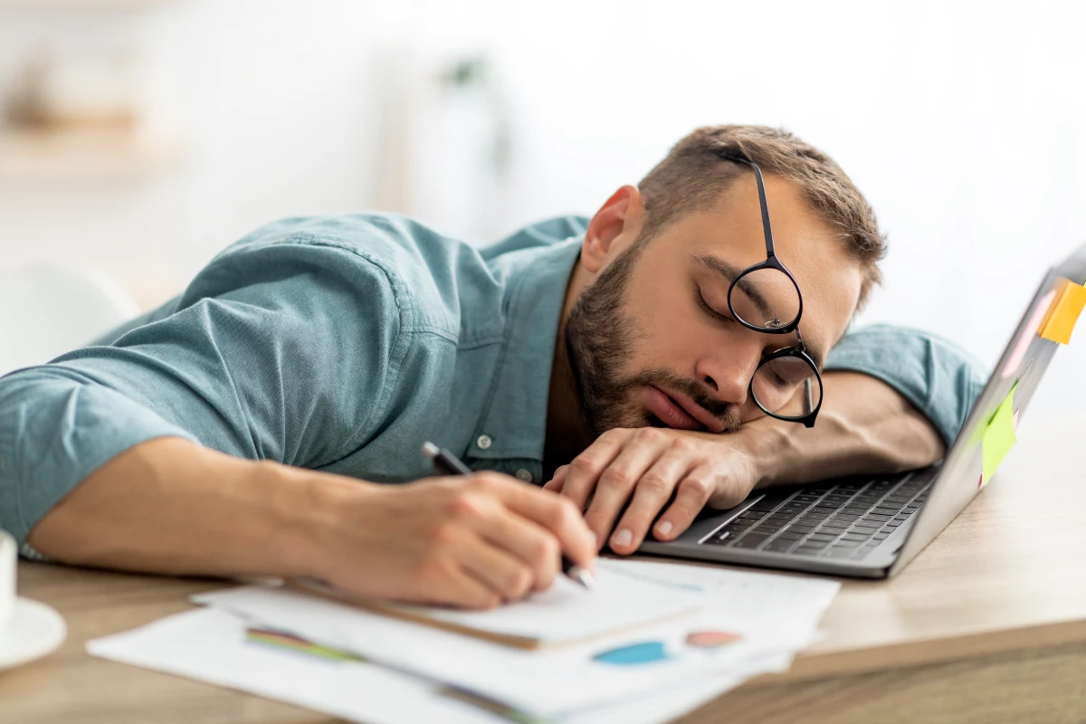 Man with glasses asleep at his desk next to a laptop, representing commonly ignored low testosterone signs such as chronic fatigue and difficulty concentrating.