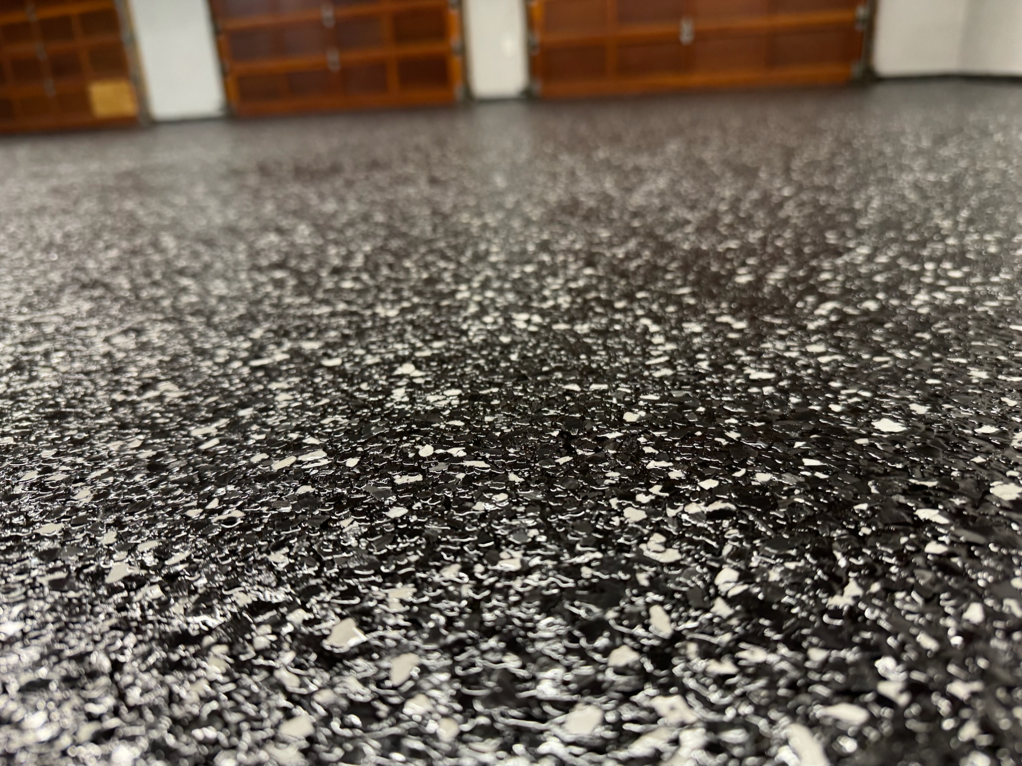 Close-up of a black professionally installed custom garage floor with white flakes and three wooden garage doors in the background in Bismarck ND