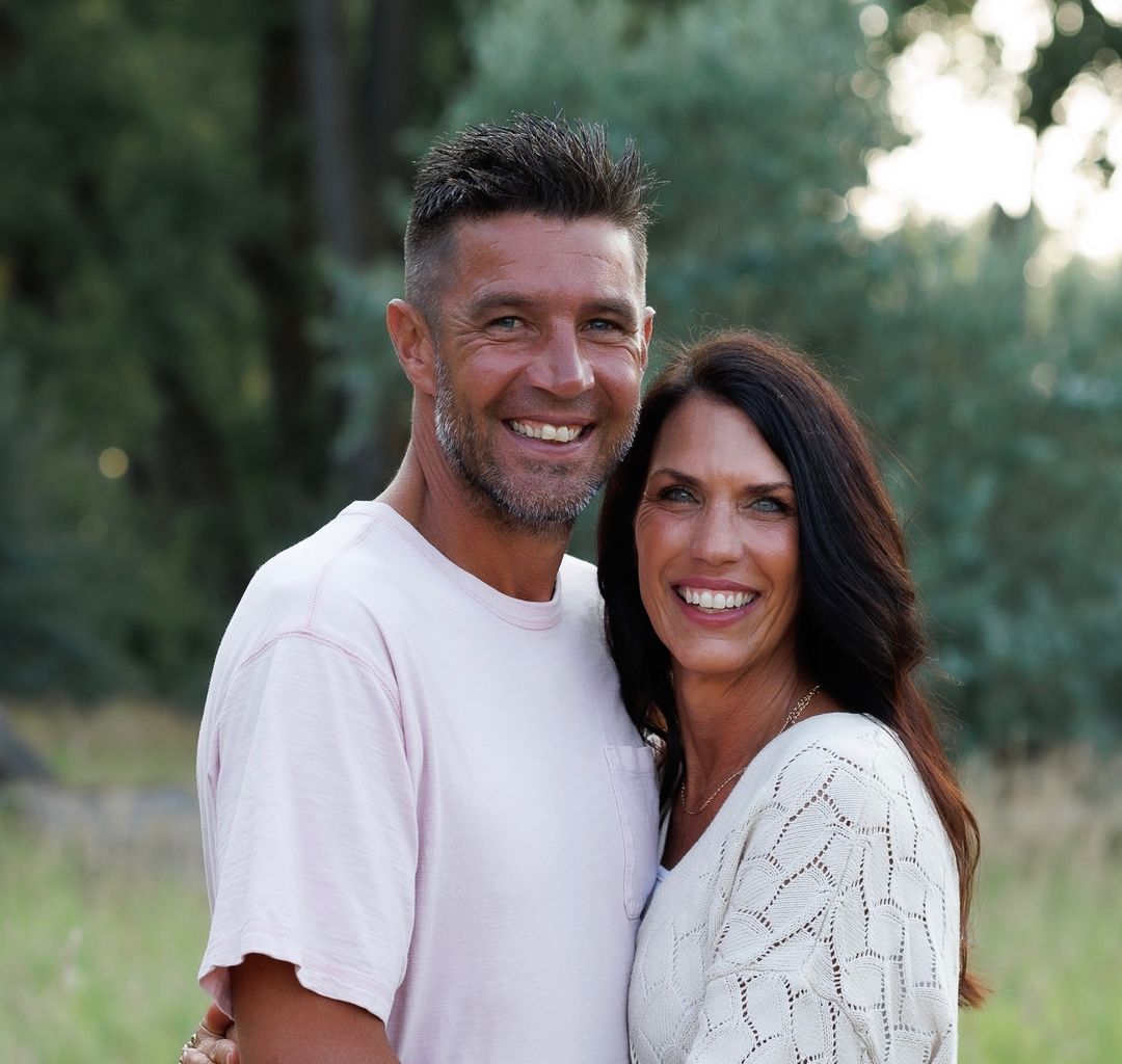 Smiling man and woman embracing outdoors with blurred trees and grass background. Brad and Nicole Stoddard. Armour Floors owners in BIsmarck ND