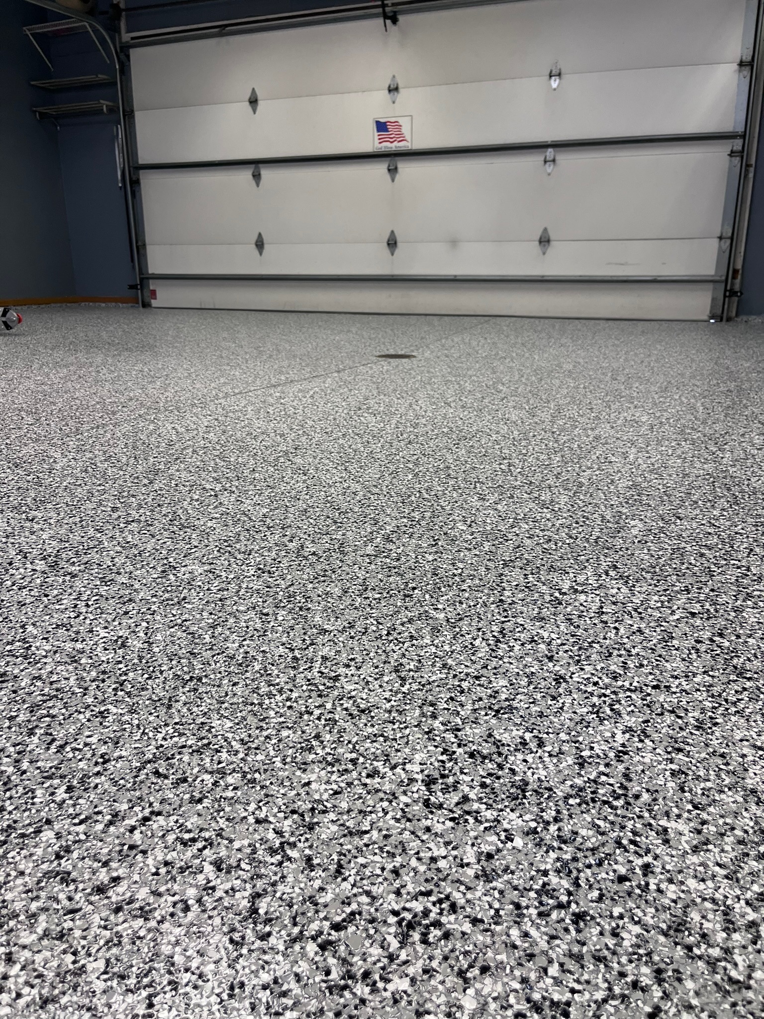 Speckled grey full broadcast flake epoxy-coated garage floor with closed white garage door and small American flag sign in BIsmarck, ND.