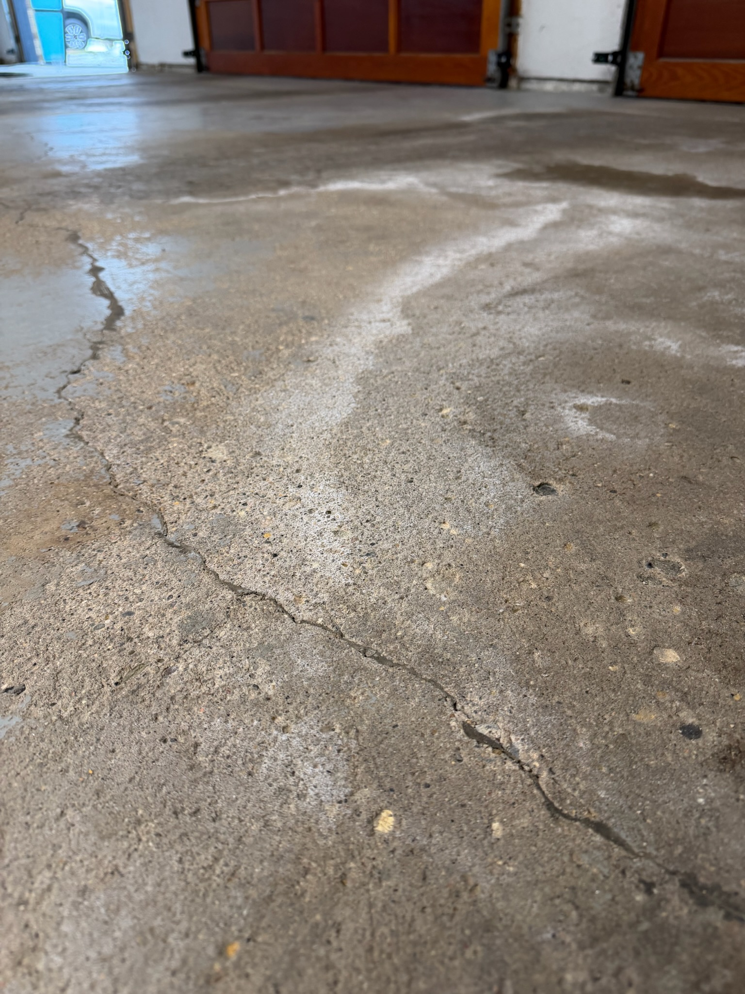 Close-up of cracked, stained concrete floor inside a garage with wooden garage doors and a car outside.