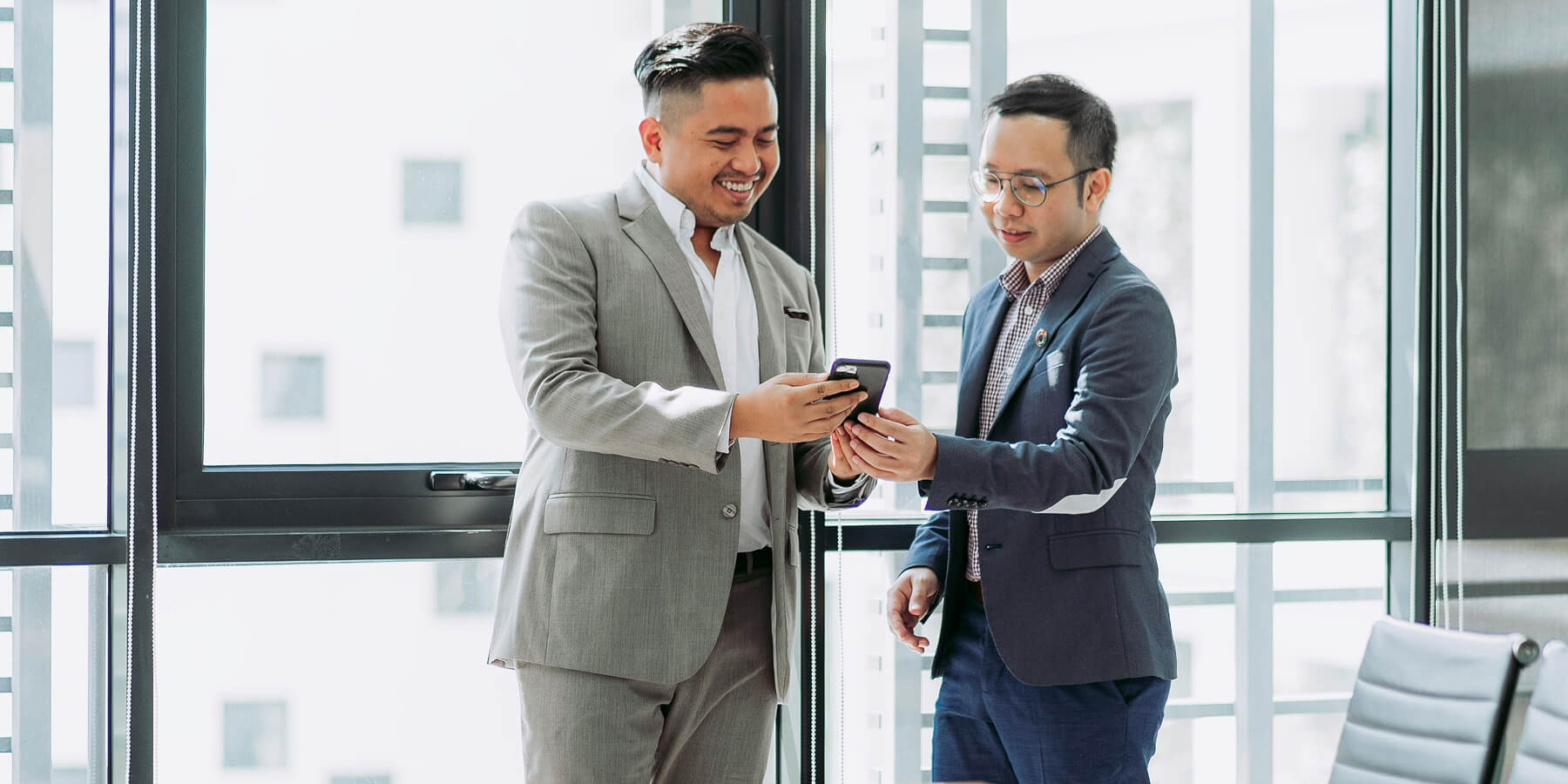 Two men in business suits smiling and looking at a smartphone together in a bright office.