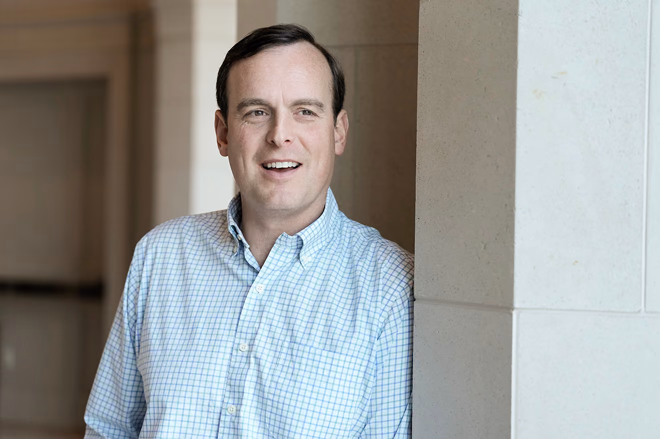 Man with short dark hair wearing a checkered shirt, leaning against a light stone wall indoors.