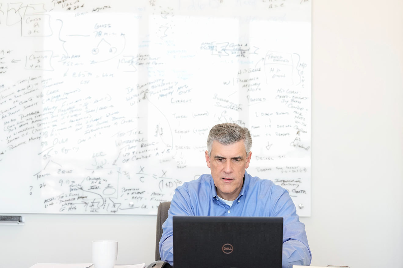 Man in blue shirt working on a laptop with a whiteboard full of handwritten notes behind him.