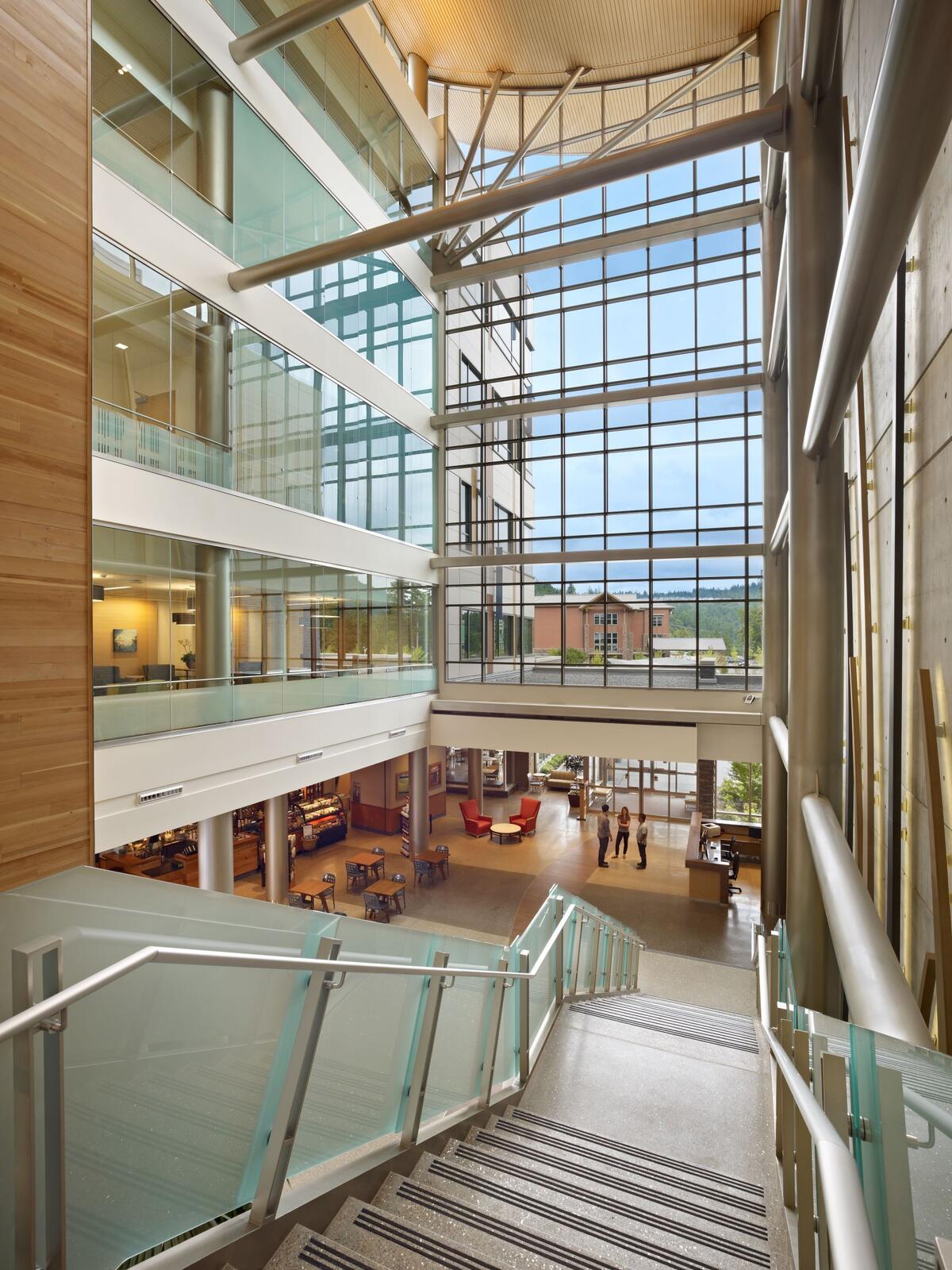 Wide modern interior stairway with glass railings overlooking a lobby with large windows and seating.
